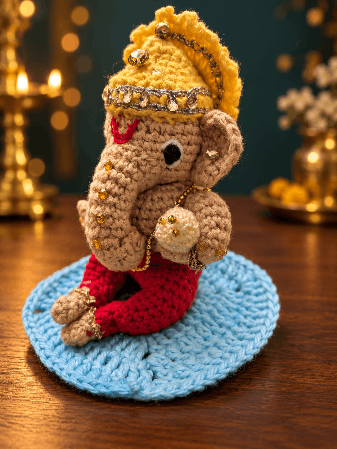 Ganesha in profile, holding his modak, on a blue crochet mat by brass lamps